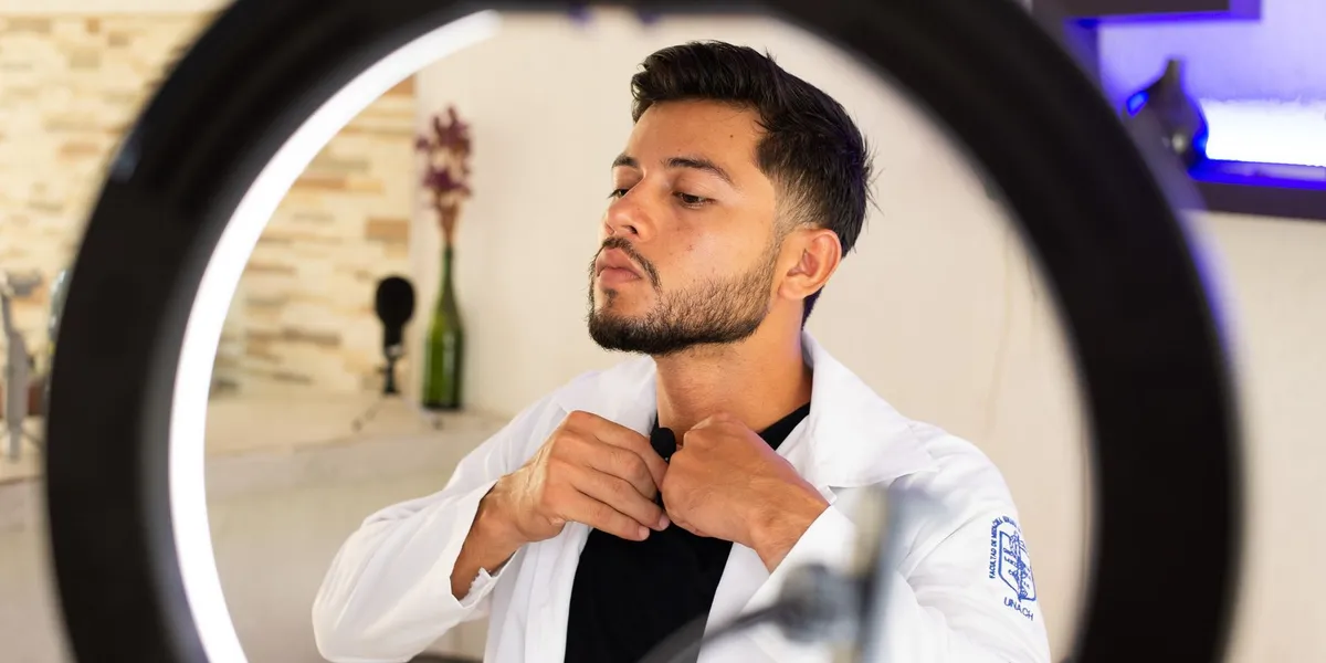 A man clips a small black lavalier microphone to the collar of his shirt, framed through the ring of a light.
