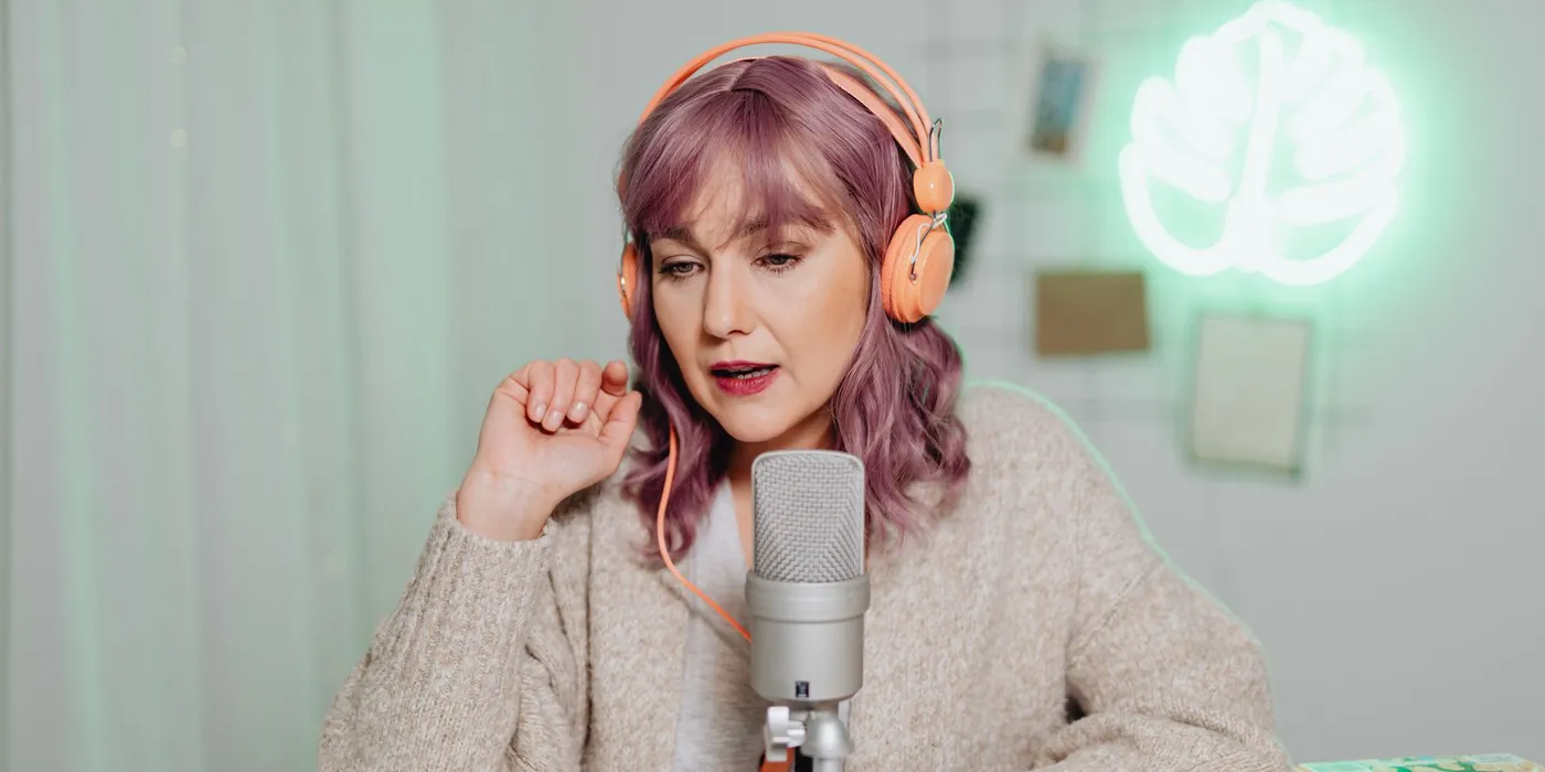 A woman wearing headphones speaks into a condenser microphone on a desk stand in a softly lit room at home.